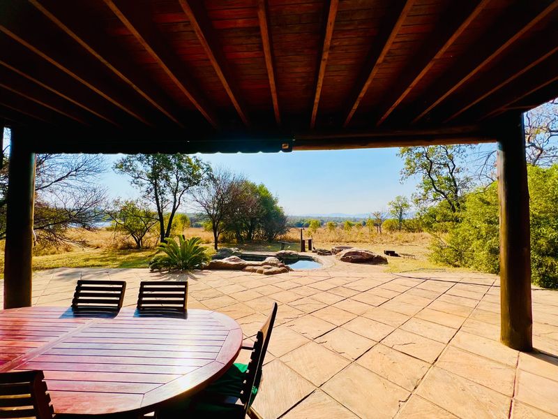 Covered patio featuring exposed timber beams and stone paving, overlooking a natural rock pool and expansive bushveld landscape.