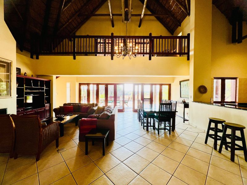 Spacious open-plan living and dining area featuring a double-volume thatched ceiling, exposed timber beams, a mezzanine level, and large folding doors leading outdoors.