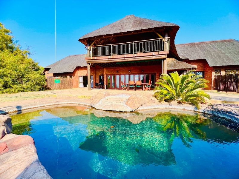 A spacious outdoor pool area featuring a natural rock-style swimming pool, a brick-built home with a thatched roof, and an upper-level balcony.