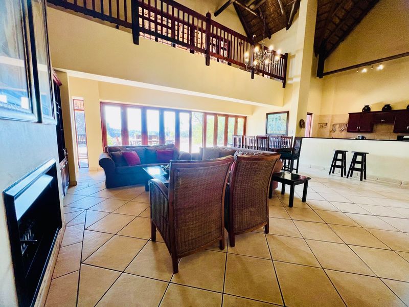 Spacious open-plan living and dining area featuring a double-volume thatched ceiling, mezzanine level, tiled floors, and large folding doors leading to the outdoors.