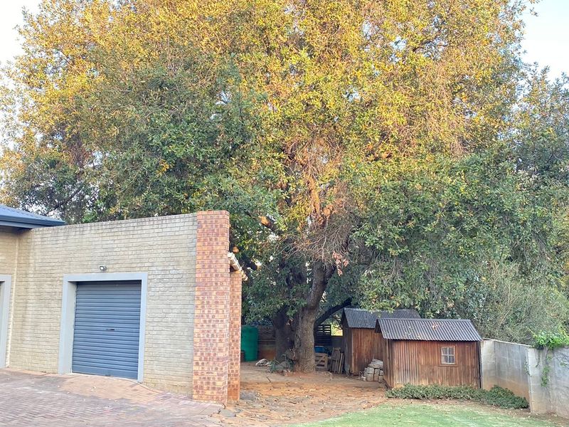 Rear exterior view featuring a double garage, brick facade, paved driveway, stone patio area, and a large mature tree with a small timber shed.