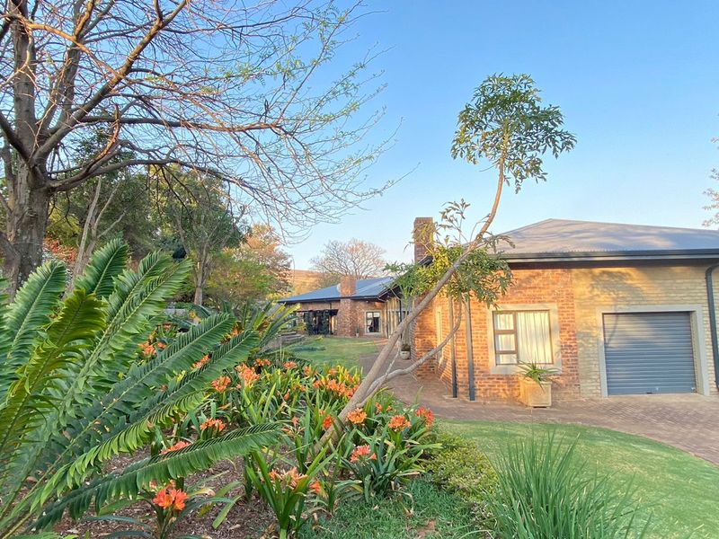 A well-maintained brick house featuring a double garage with roller doors, a paved driveway, and a lush, established garden with mature trees.