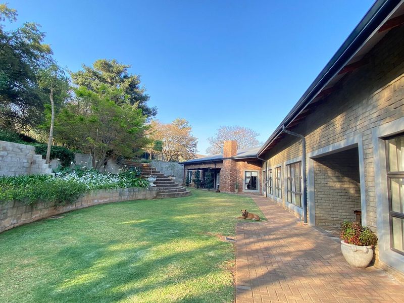 A spacious rear garden featuring a well-maintained lawn, brick-paved walkway, stone staircase, and a charming brick facade with large aluminum-framed windows.