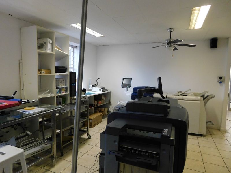 A functional office or workspace featuring tiled flooring, built-in shelving, and overhead fluorescent lighting, currently utilized for printing and equipment storage.