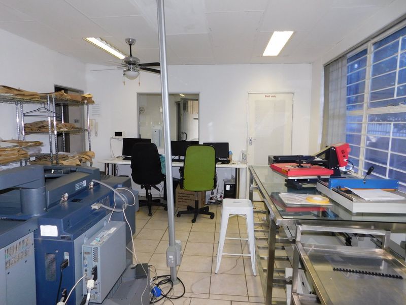 Functional office space featuring tiled flooring, fluorescent lighting, and security bars on windows, currently utilized as a workspace with various equipment.