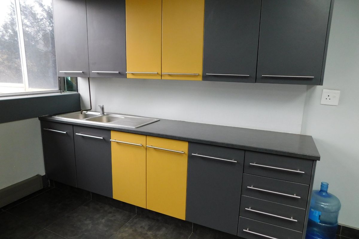 A modern office kitchenette featuring contrasting grey and yellow cabinetry, dark stone-look countertops, and a stainless steel sink.
