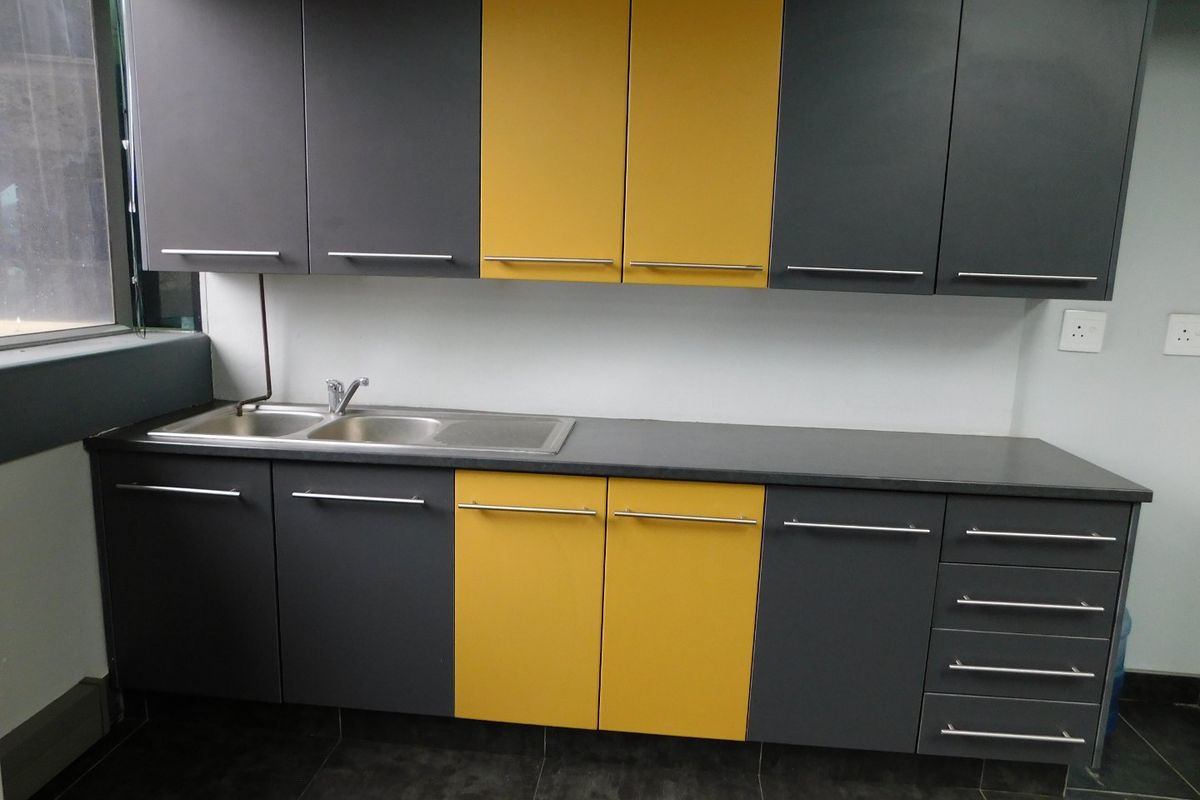 A modern office kitchenette featuring contrasting grey and yellow cabinetry, a stainless steel sink, and dark tiled flooring.