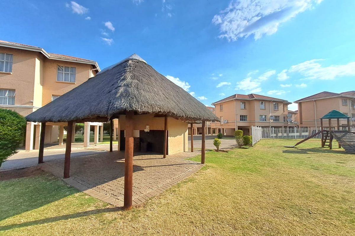 Communal outdoor area featuring a thatched lapa with paved flooring, set within a well-maintained garden with children's play equipment.