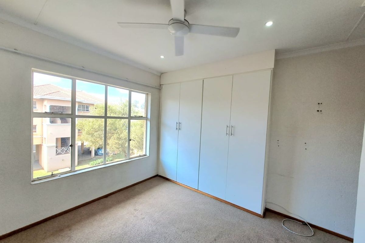 A bright, empty bedroom featuring built-in cupboards, a ceiling fan, recessed lighting, and large windows overlooking the complex grounds.