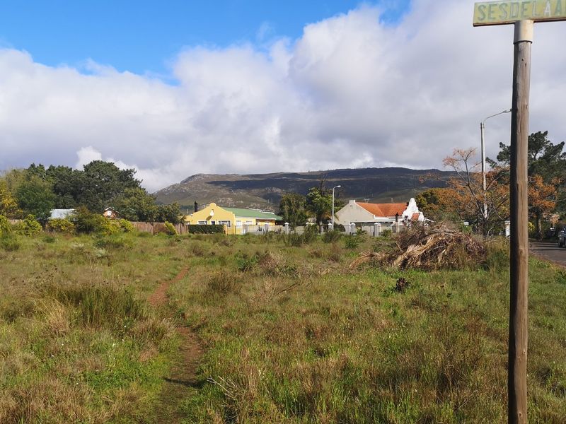 A view of a vacant residential plot featuring natural vegetation, with surrounding neighborhood houses and mountain views in the background.