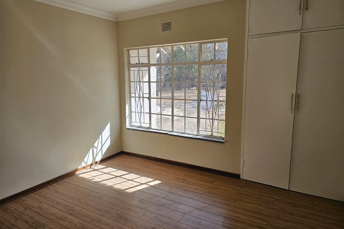 A bright, empty bedroom featuring wood-look laminate flooring, built-in cupboards, and a large window with security bars.