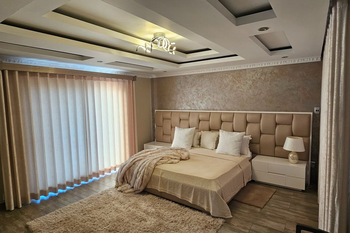 Spacious bedroom featuring a decorative tray ceiling with recessed lighting, a large built-in upholstered headboard, and tiled flooring.