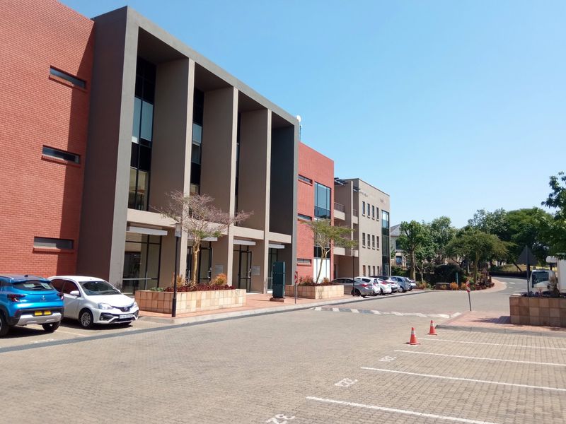 Modern office building exterior featuring a red brick and concrete facade with large glass windows and an adjacent paved parking area.