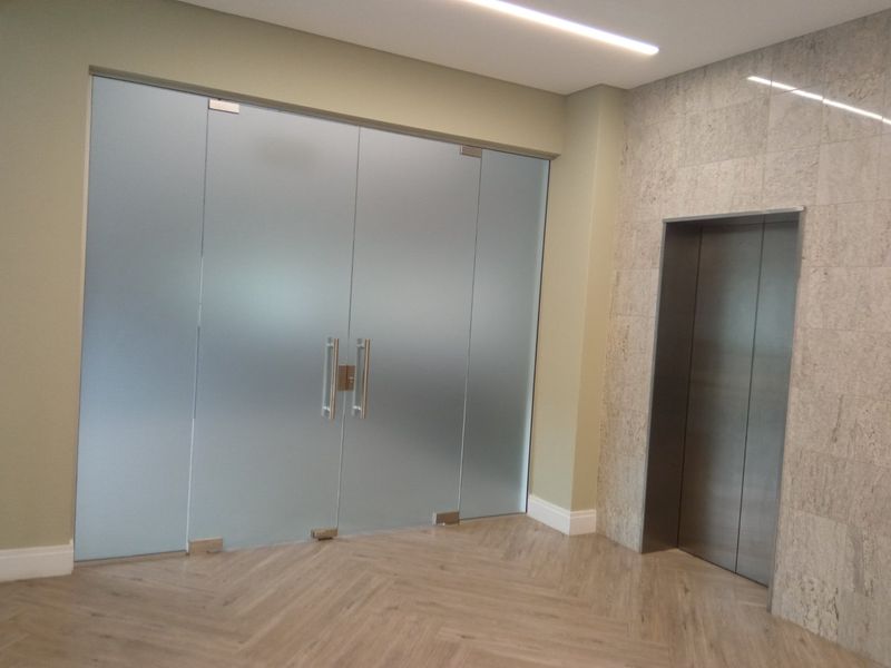 Modern office lobby area featuring frosted glass double doors, a stainless steel elevator, and elegant herringbone flooring with stone tile accents.