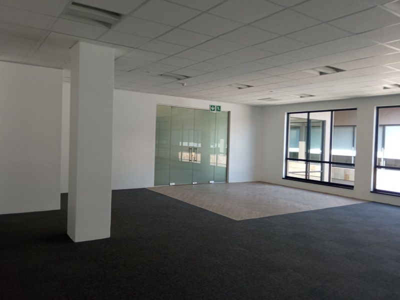 A spacious, vacant open-plan office area featuring a mix of carpet and herringbone flooring, a suspended ceiling, and a modern glass partition door.