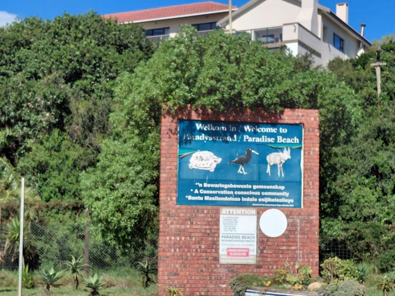 A brick entrance sign for the Paradise Beach community, featuring local wildlife illustrations and neighborhood watch information, set against lush greenery.