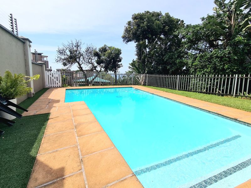 A well-maintained outdoor swimming pool area featuring paved surrounds, secure perimeter fencing, and established garden greenery.