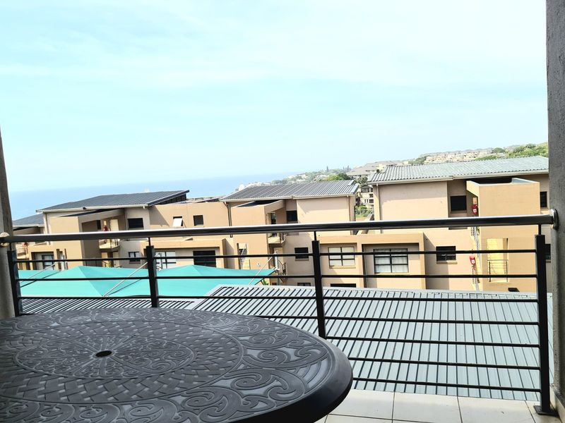 A balcony featuring a stainless steel balustrade and tiled flooring, offering an elevated view of the surrounding residential complex and distant coastline.