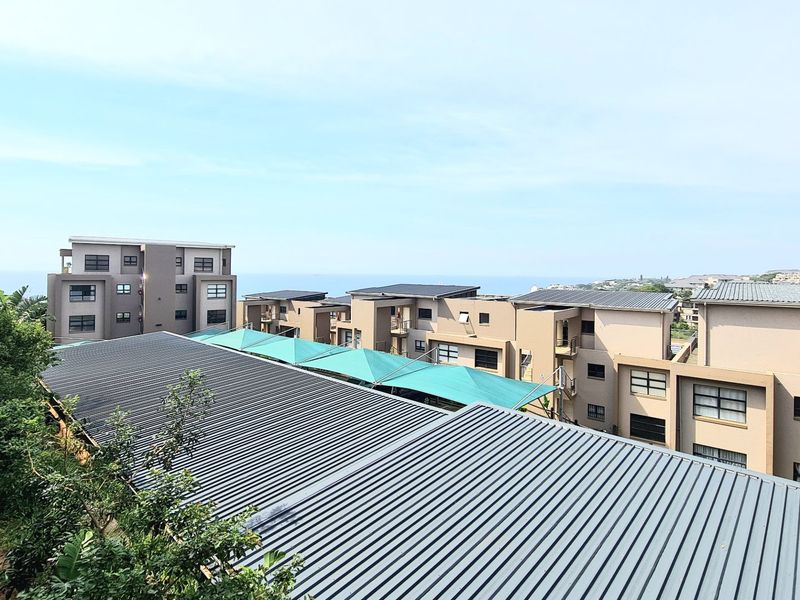 Elevated view overlooking the apartment complex, featuring modern building architecture, metal roofing, and shaded parking structures with a distant sea view.