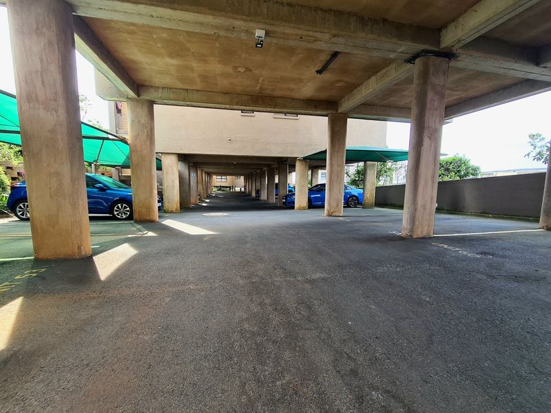 Covered parking area featuring concrete structural pillars and asphalt surfacing, providing sheltered vehicle storage for the flat complex.