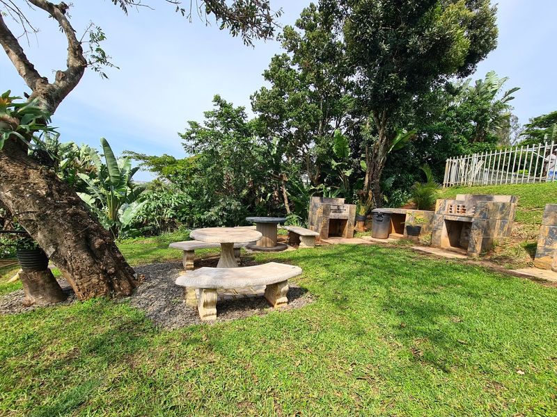 A landscaped garden area featuring multiple built-in stone braai stations, stone seating, and mature trees, providing an established outdoor entertainment space.