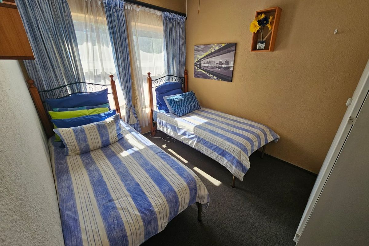 A compact bedroom featuring neutral textured walls, dark carpeting, and large windows with curtain treatments, providing a functional space for sleeping.