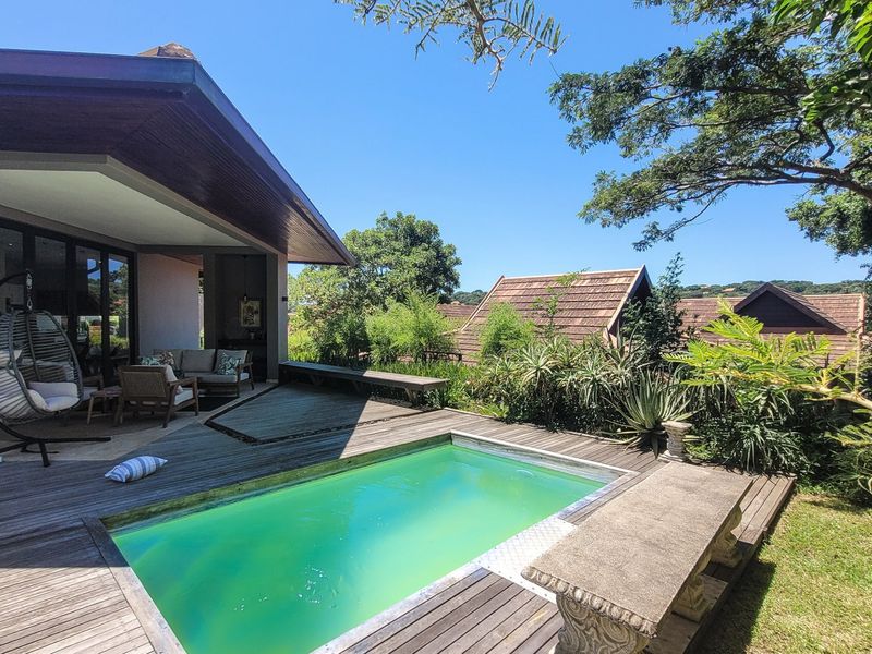 Outdoor pool area featuring timber decking, a covered patio space, and a stone bench, set within a landscaped garden environment.