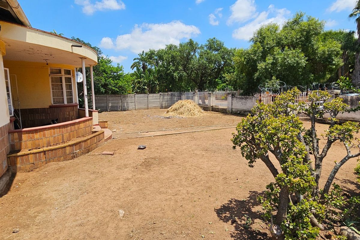 A spacious rear garden area featuring a covered patio with brick detailing, mature trees, and a perimeter boundary wall.