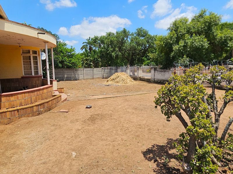 A spacious rear garden area featuring a covered patio with brick detailing, mature trees, and a perimeter boundary wall.