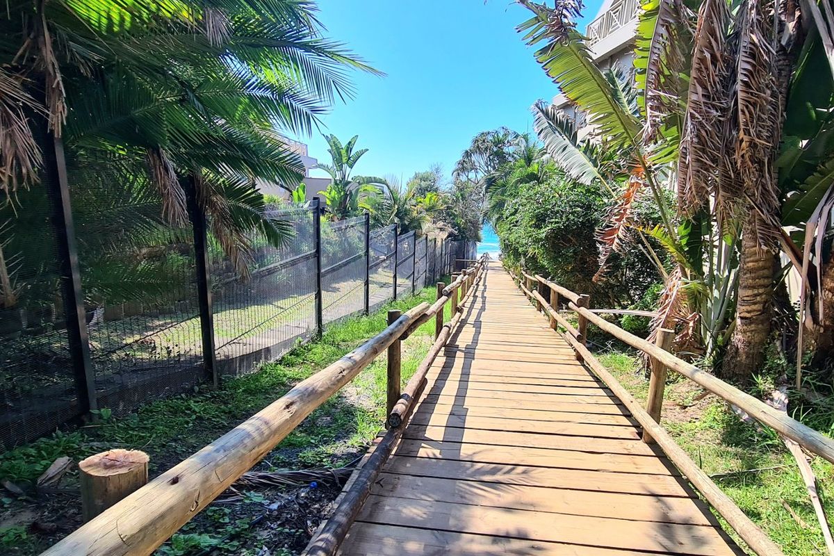A wooden boardwalk pathway leading towards the beach, flanked by mature palm trees and perimeter fencing.