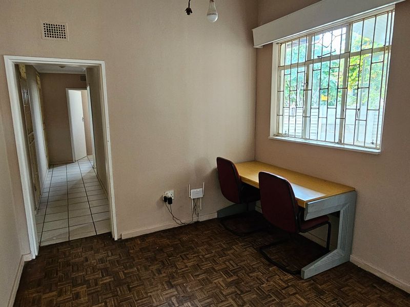 A functional office space featuring parquet flooring, a large window with security bars, and direct access to the hallway.