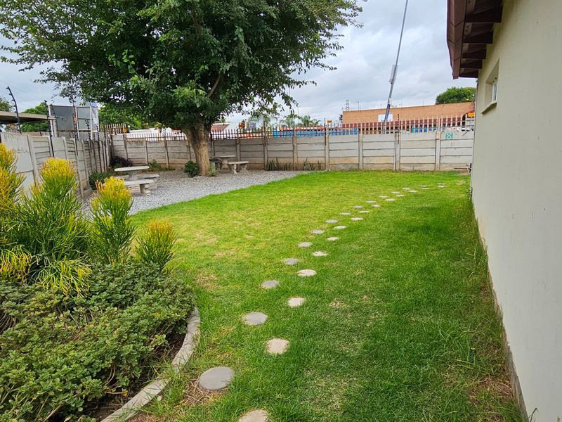 A private rear garden featuring a lawn, stone pathway, established tree, and concrete boundary wall with security fencing.
