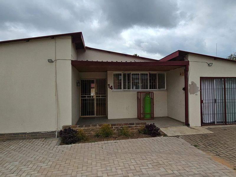 Front elevation of a single-story house featuring a paved driveway, covered entrance porch, security gates, and a low-maintenance garden area.