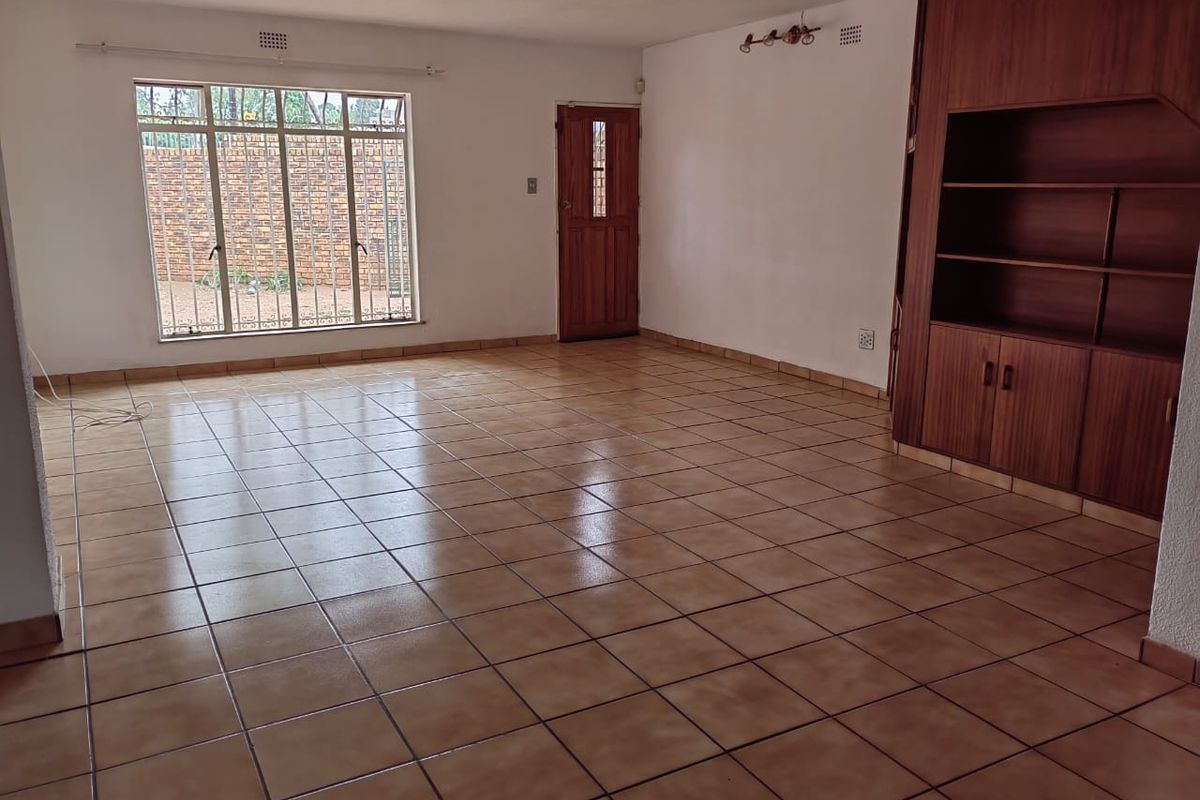 Spacious, empty living area featuring neutral tiled flooring, built-in wooden cabinetry, and a secure entrance door with natural light from a large window.
