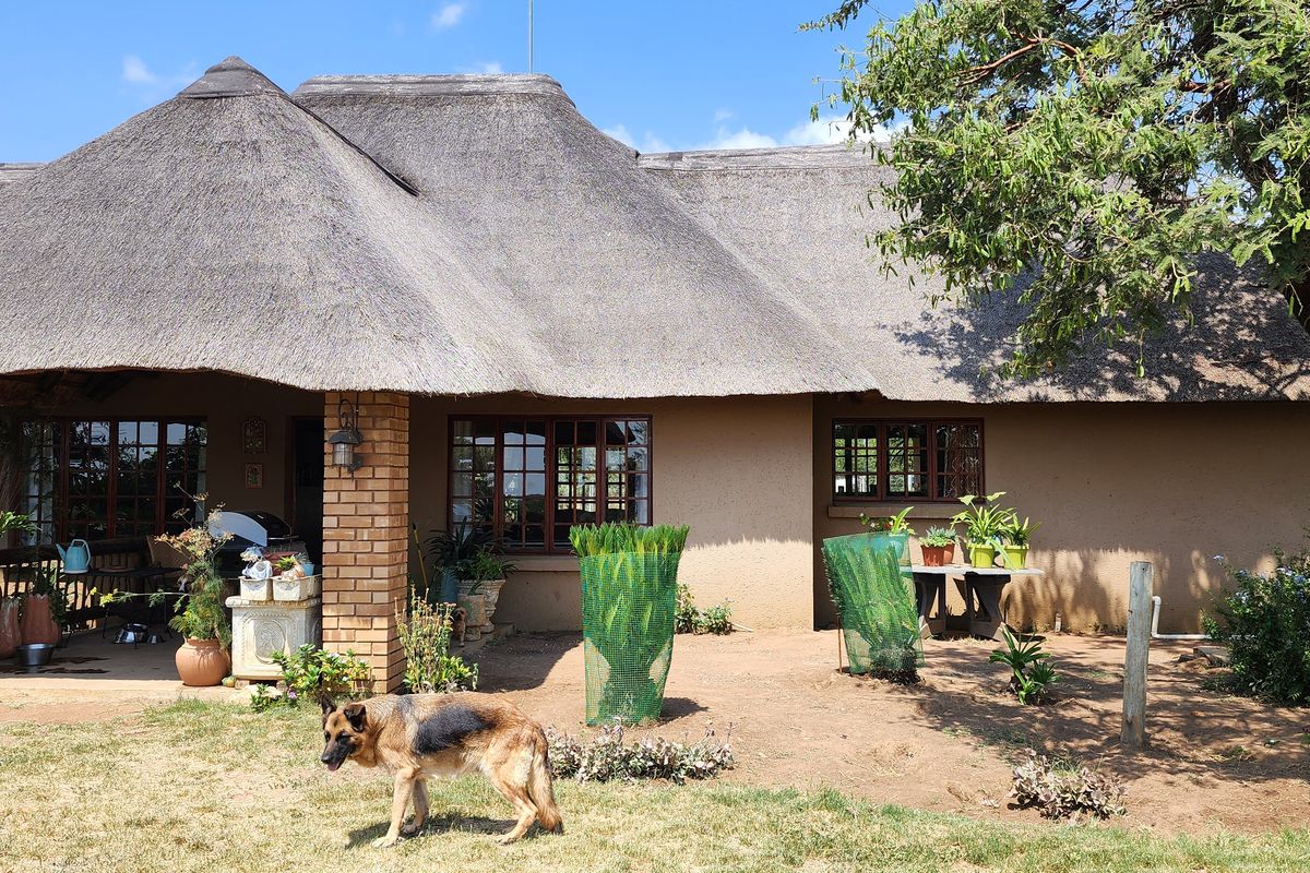 A traditional farmhouse exterior featuring a classic thatched roof, brick support pillars, and a covered patio area overlooking the surrounding garden.