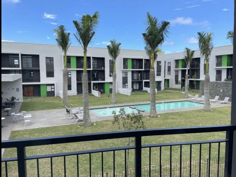 View of a modern residential complex featuring a communal swimming pool, landscaped garden, and paved patio area, viewed from an elevated balcony.