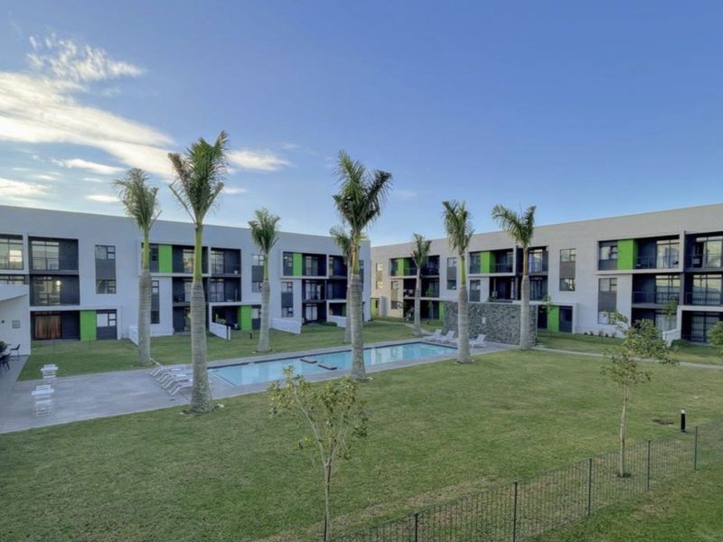 A well-maintained communal pool area featuring a swimming pool, landscaped garden, and paved patio, surrounded by modern multi-story apartment buildings.