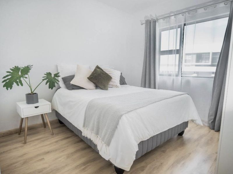 A bright and well-maintained bedroom featuring light-toned laminate flooring and a large window with modern aluminium frames.