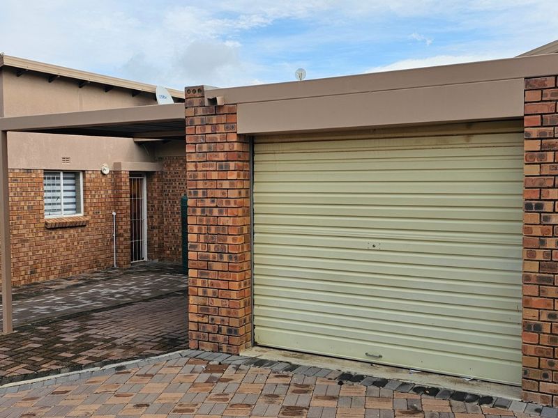 Exterior view of a townhouse featuring a brick facade, a single garage with a roll-up door, and a covered carport area with paved driveway.