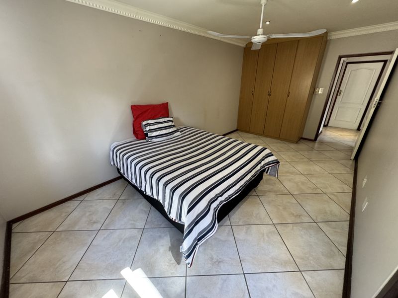 A bedroom featuring tiled flooring, built-in wooden cupboards, and a ceiling fan, offering a functional and neutral space.