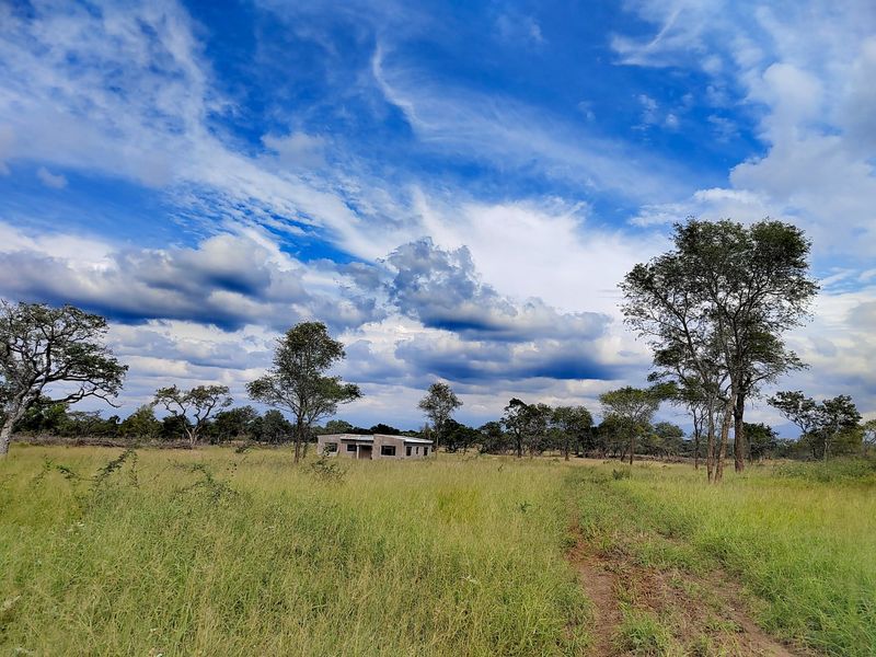 A wide-angle view of a rural farm property featuring an unfinished concrete block structure set within a large, grassy landscape.
