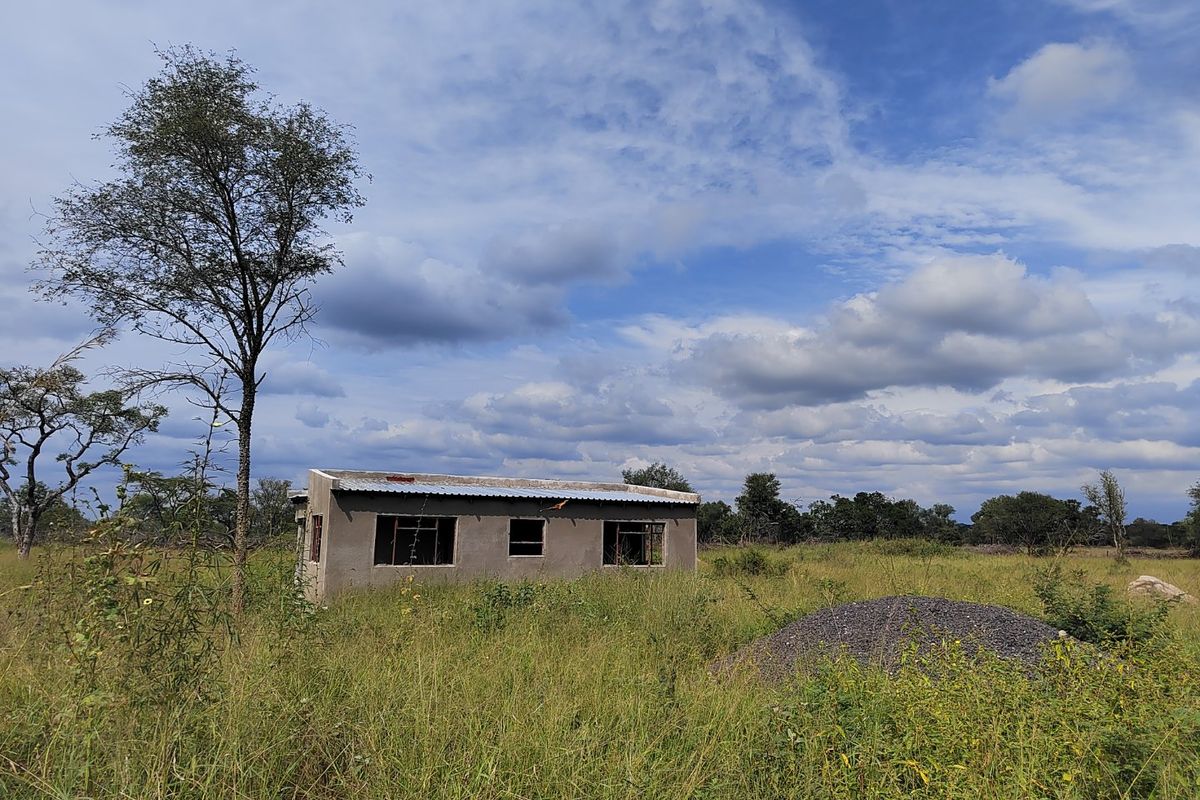 A small, unfinished masonry structure situated on a large, overgrown rural plot, featuring a corrugated metal roof and open window frames.
