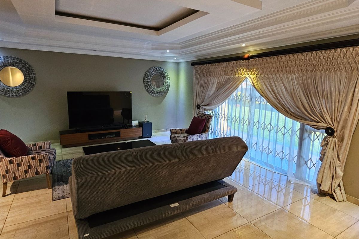 A well-maintained living room featuring polished floor tiles, a recessed tray ceiling with integrated downlighting, and a large sliding door with security gate.