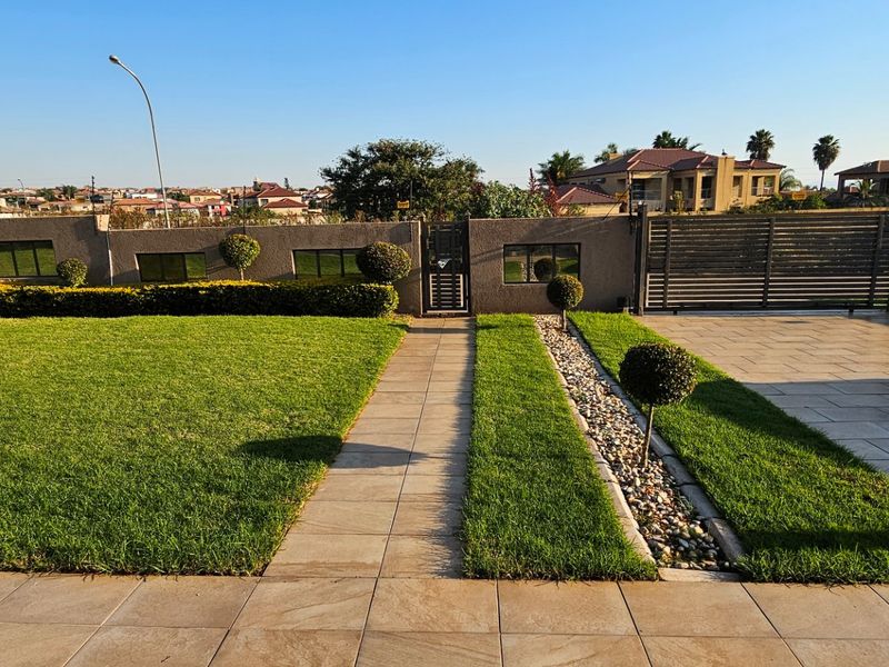 A well-maintained front garden featuring a paved walkway, manicured lawn, decorative stone border, and a secure perimeter wall with a gated entrance.