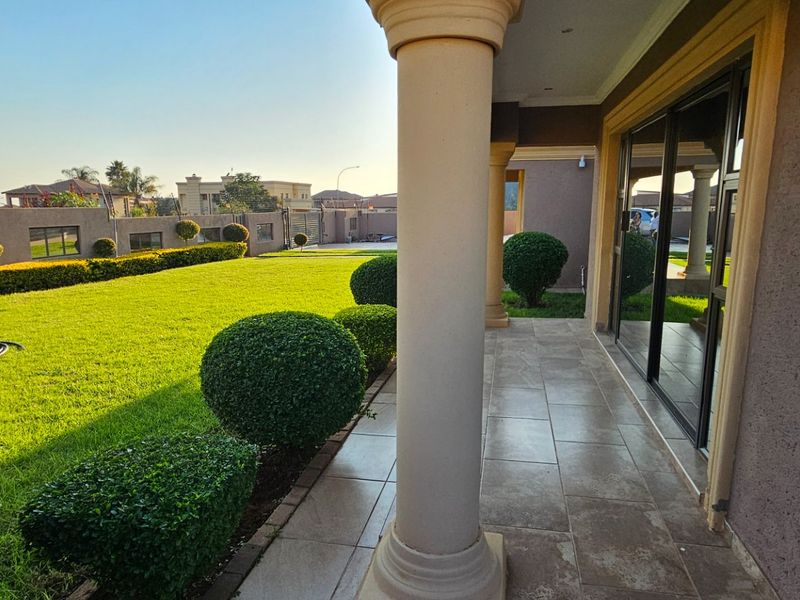 A spacious covered patio featuring tiled flooring and structural columns, overlooking a well-maintained garden with manicured shrubs and a perimeter wall.