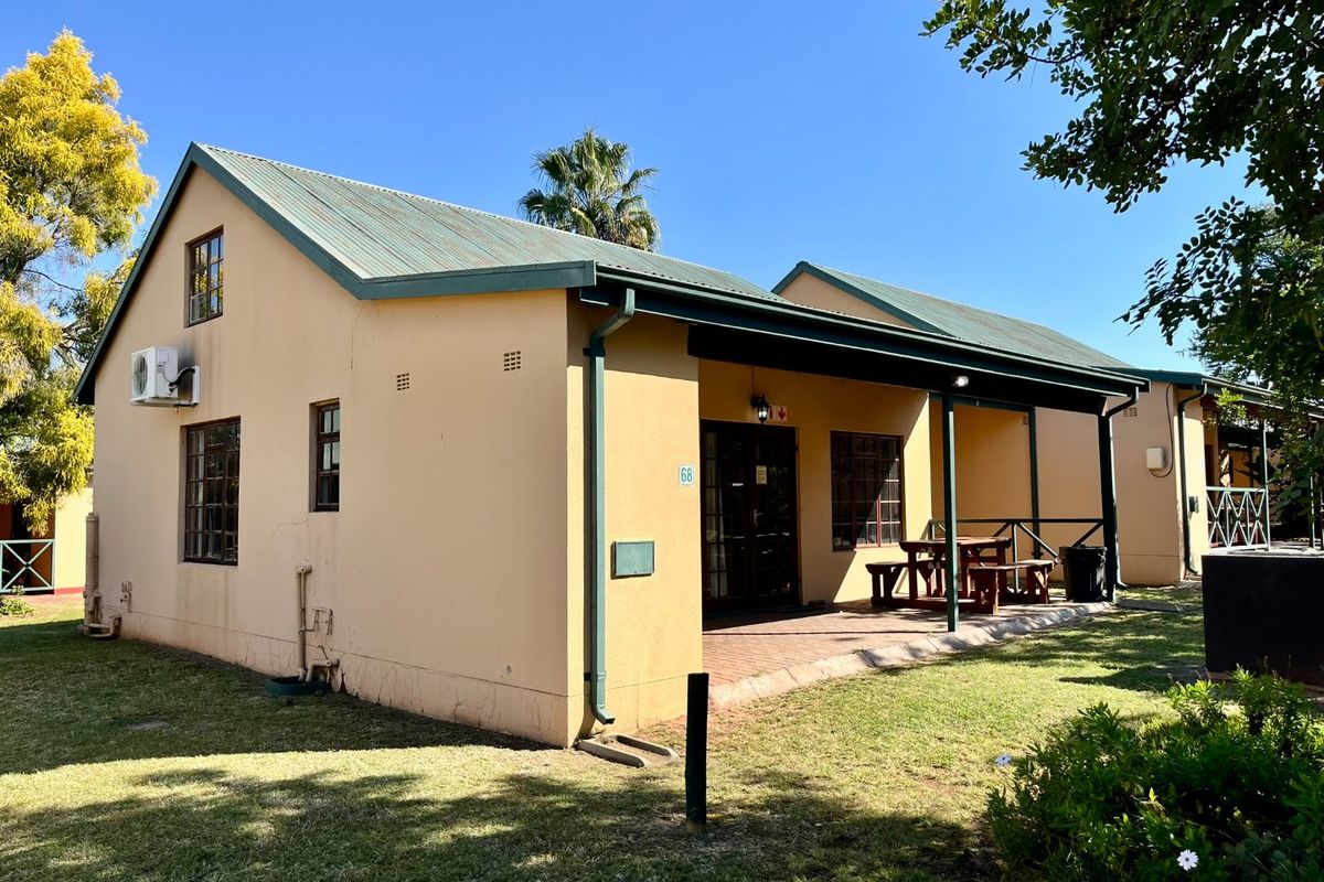 Rear view of a single-story flat featuring a covered patio, brick paving, and a pitched metal roof set within a landscaped garden area.