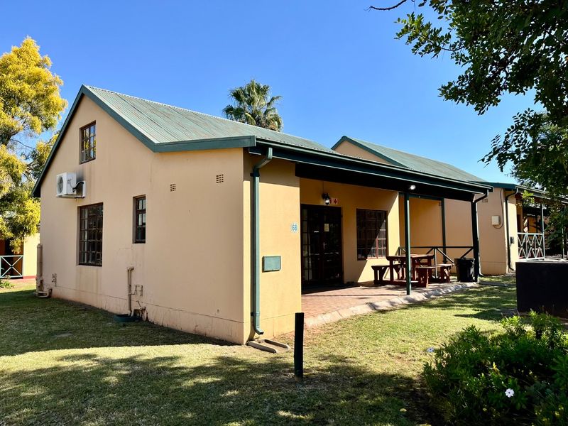 Rear view of a single-story flat featuring a covered patio, brick paving, and a pitched metal roof set within a landscaped garden area.
