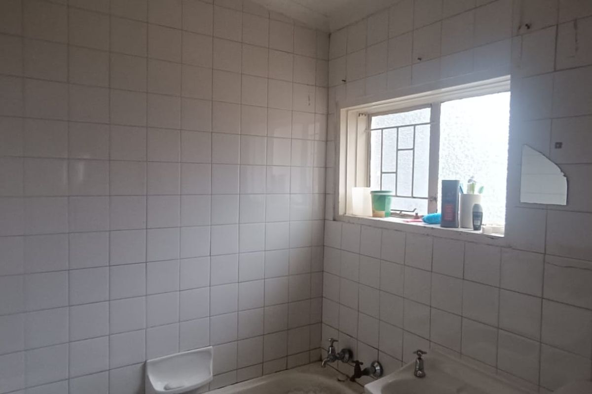 A dated bathroom featuring white wall tiles, a built-in bathtub, and a window with security bars, requiring cosmetic updates and maintenance.