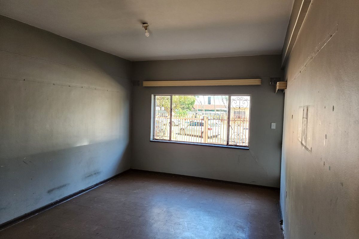 An empty, dated living room featuring a large window with security bars, neutral wall paint, and original flooring, ready for renovation.