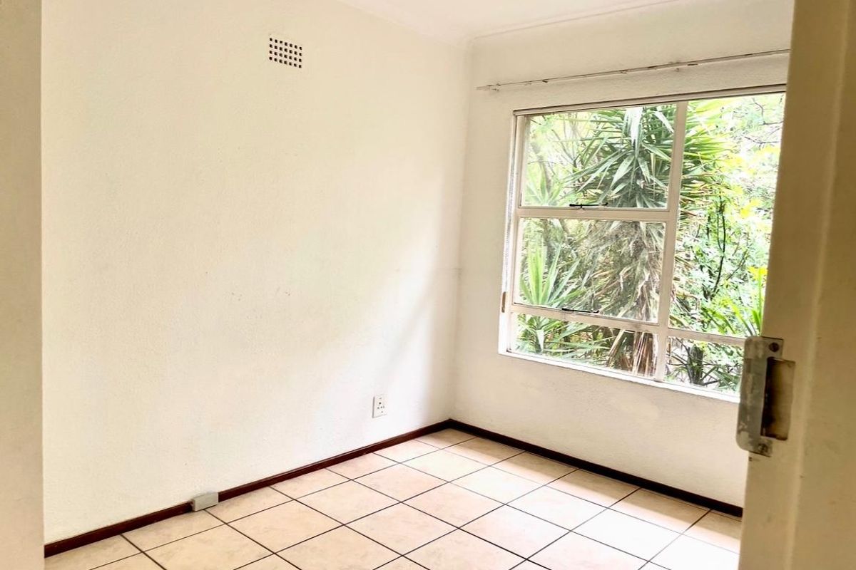 A bright, empty bedroom featuring tiled flooring, a large window with garden views, and a built-in ventilation vent.